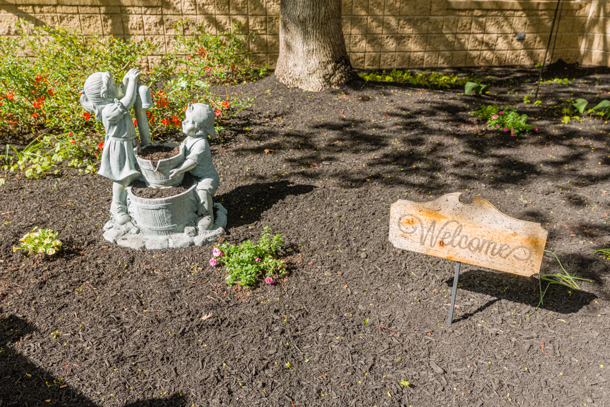 A garden area with a statue of two children playing with a bucket and a watering can, surrounded by mulch and small flowering plants. There is a wooden sign with the word 'Welcome' written on it, and a tree trunk and brick wall in the background.