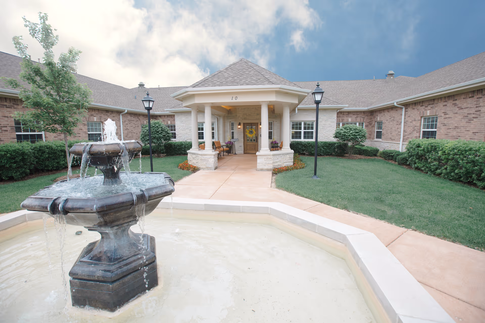 Front entrance of a single-story brick building with a fountain in the foreground and a covered portico.
