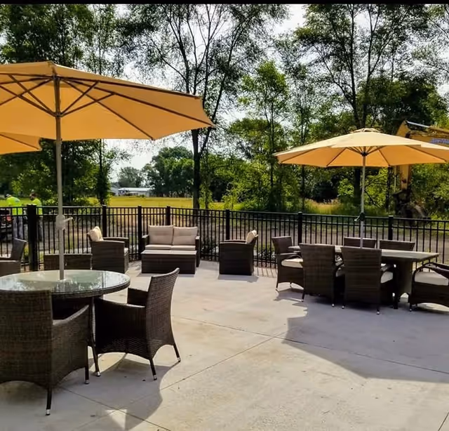 Outdoor patio area with multiple round and rectangular tables surrounded by wicker chairs. Large yellow umbrellas provide shade over the tables. In the background, there are trees and a black metal fence enclosing the patio.