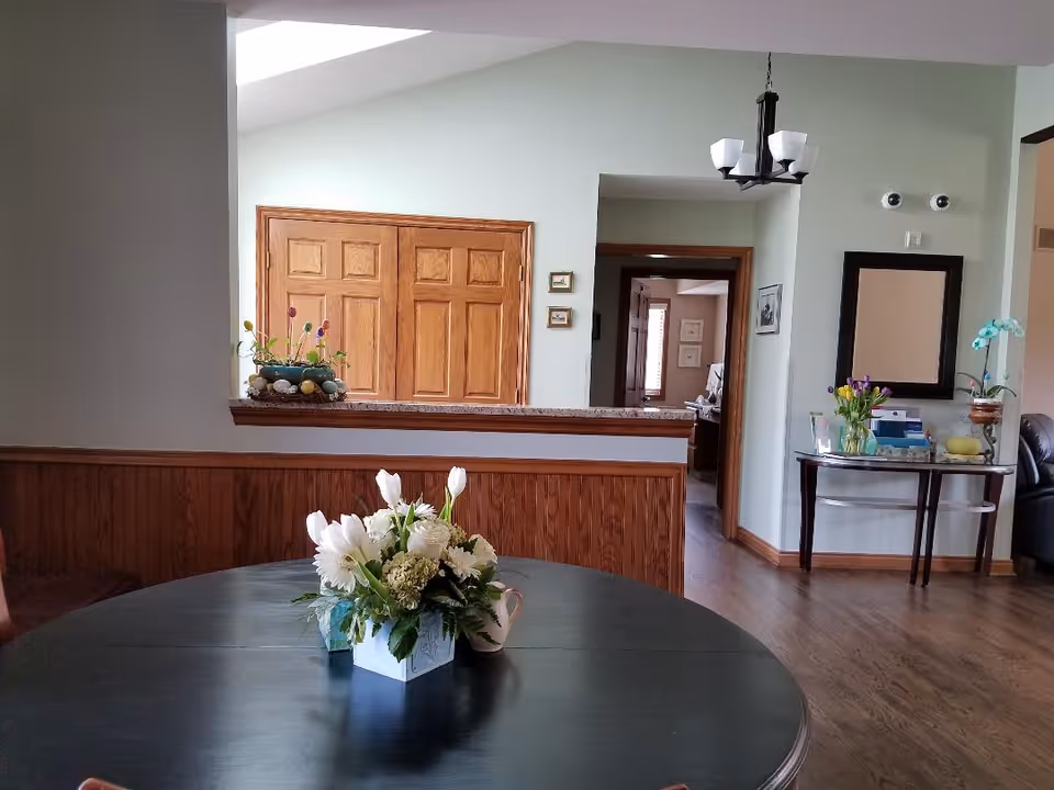 Interior view of a room in an assisted living facility featuring a round dark wooden table with a floral centerpiece. The room has light green walls, wooden flooring, and wooden trim. There is a half wall with a granite countertop, a wooden double door, a small table with flowers and a mirror on the right wall, and a chandelier hanging from the ceiling.