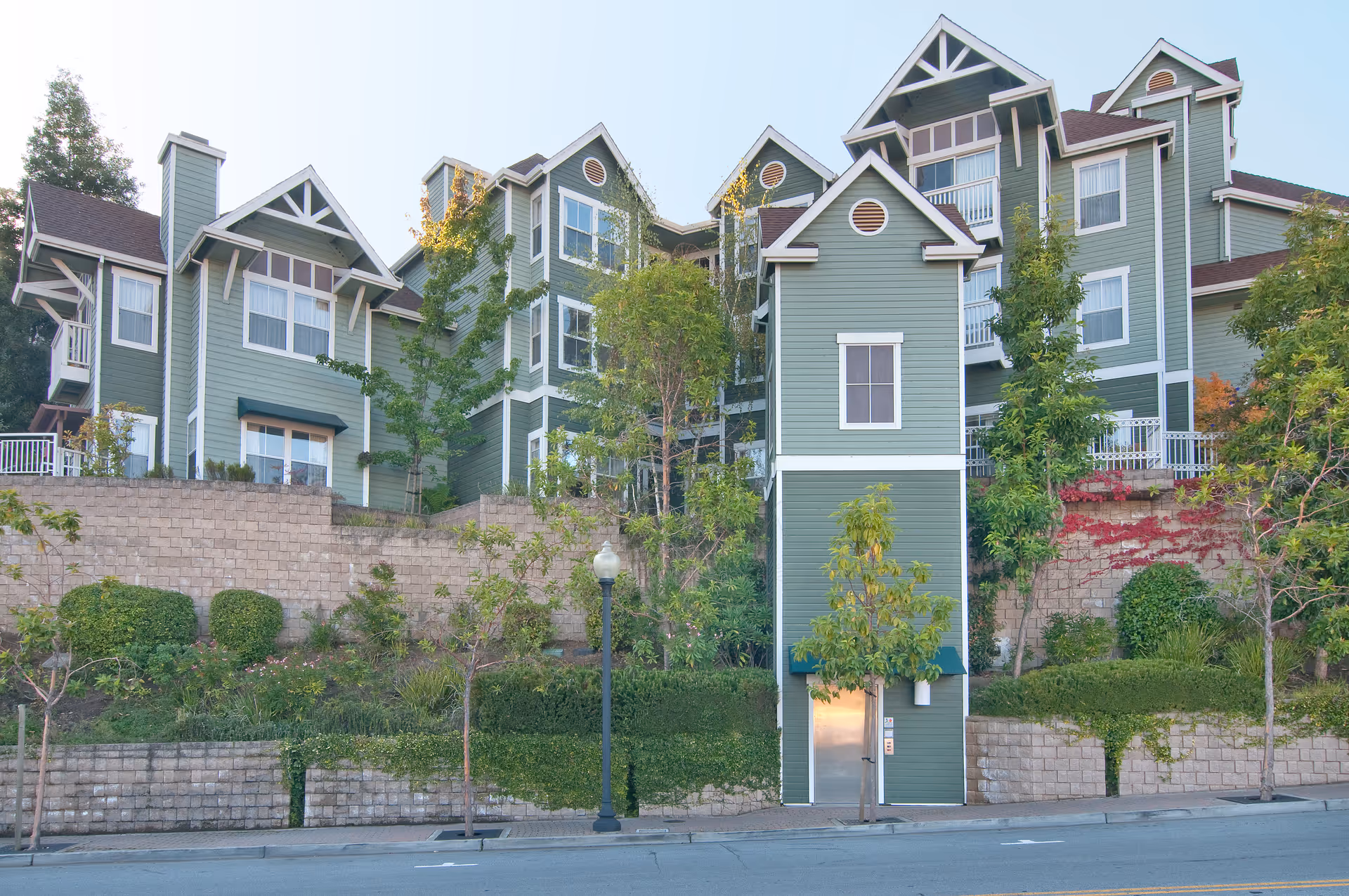 Front exterior of a green multi-story residential building with terraced stone retaining walls, landscaping, and a small elevator tower at street level.