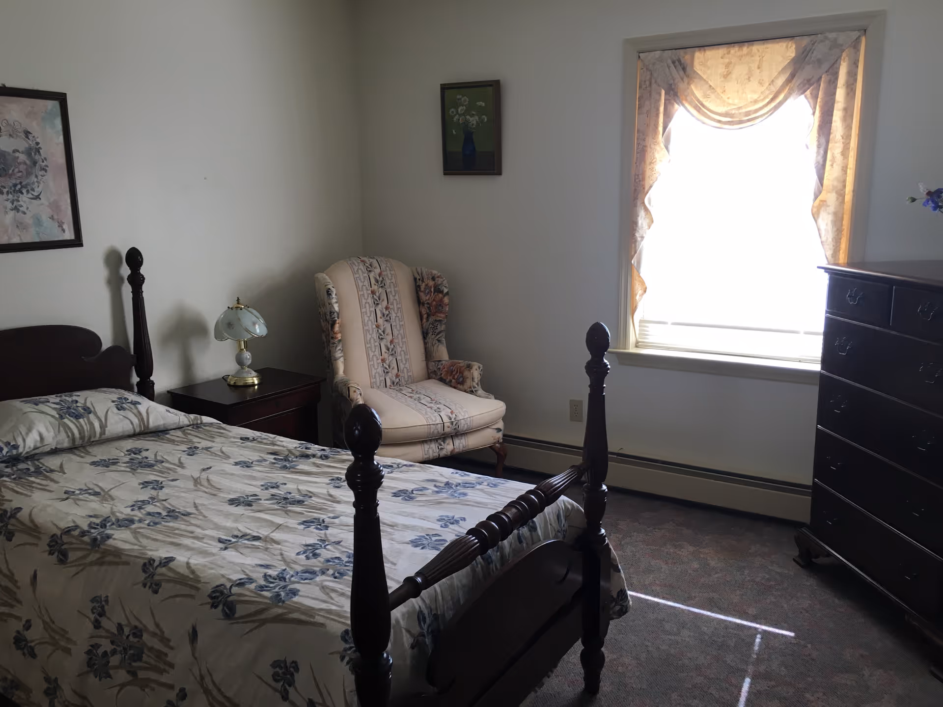 Sunlit bedroom with a wooden four-poster bed with floral bedding, an upholstered armchair, nightstand with lamp, and a tall dresser by the window.
