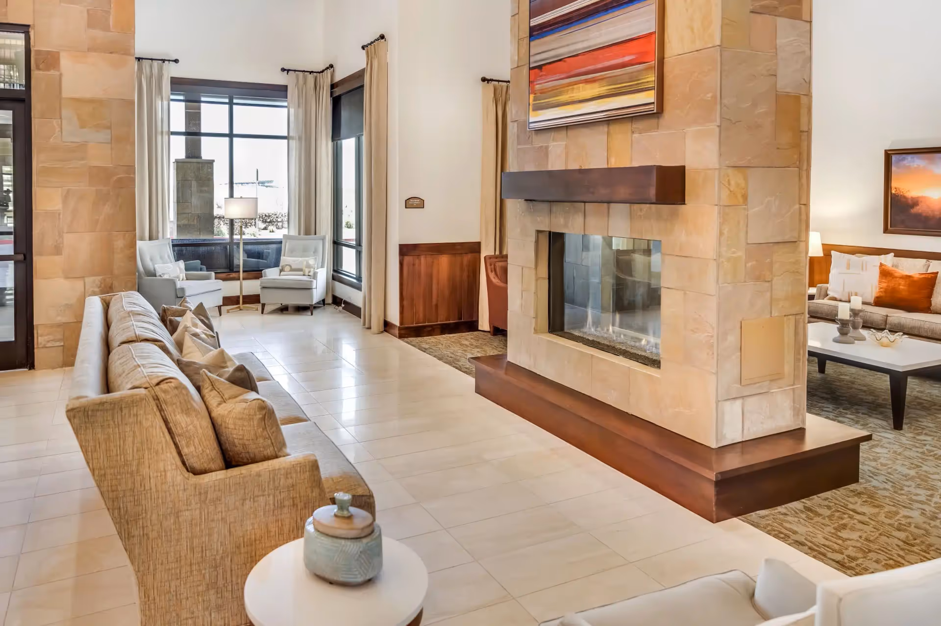 Bright modern communal living room with sofas, armchairs, and a central stone fireplace.