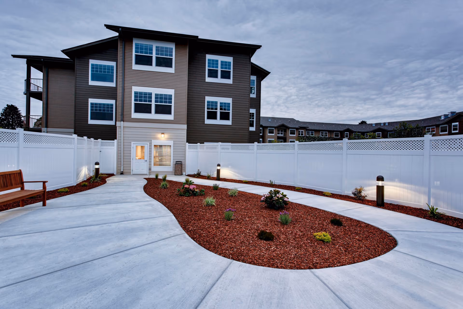 Outdoor courtyard area at Sea View Senior Living Community with a curved concrete walkway surrounding a landscaped garden bed with small plants and mulch, white privacy fencing, a wooden bench on the left, and a multi-story building in the background under a cloudy sky.