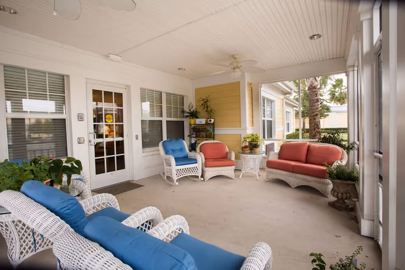 A covered outdoor patio area at The Brennity at Melbourne Senior Living featuring white wicker furniture with blue and red cushions, potted plants, ceiling fans, and large windows and doors leading inside.