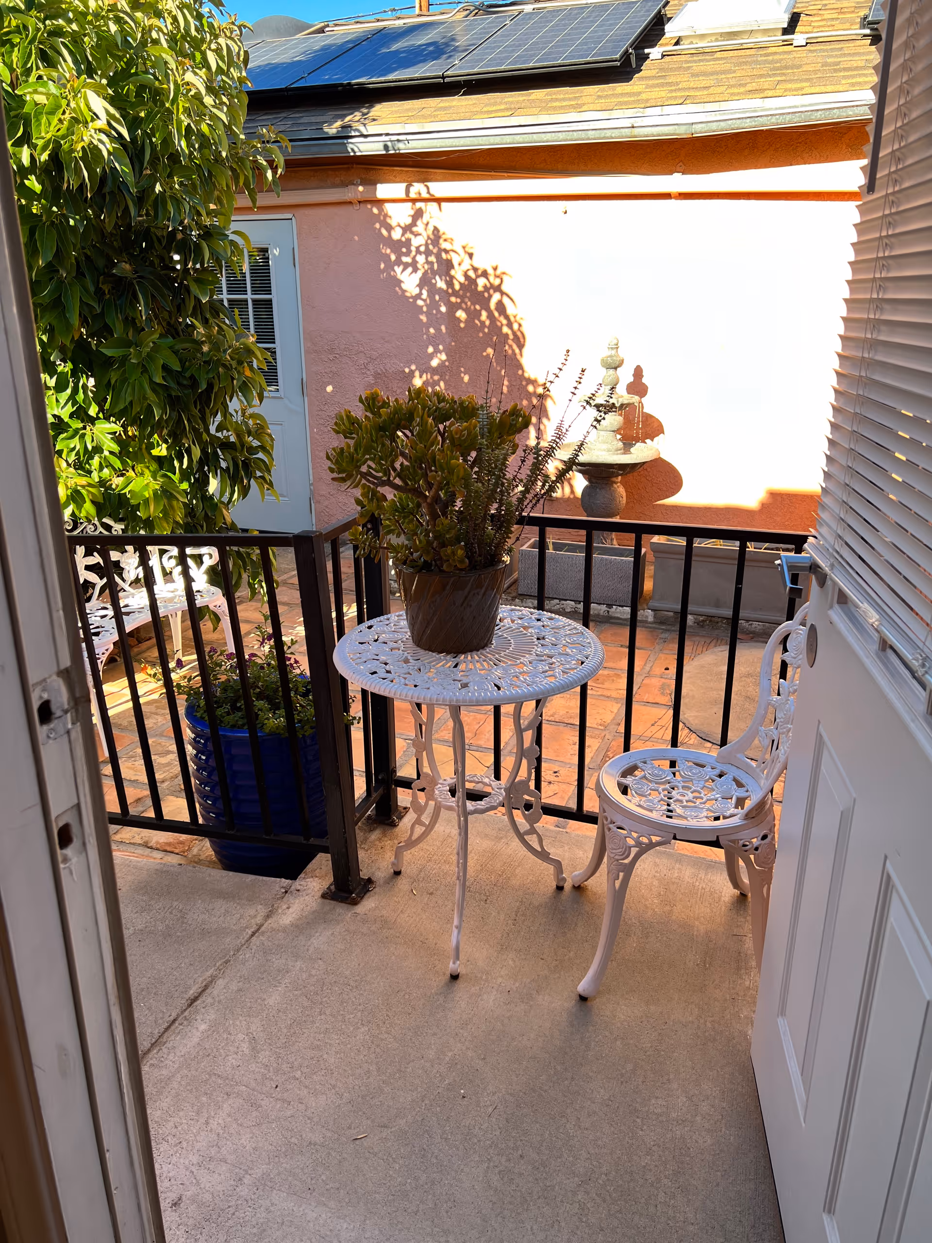 View from an open door onto a small outdoor patio area with a white metal table and chair set. A potted plant sits on the table. There is a black metal railing around the patio, a large green leafy plant in a blue pot, and a pink wall with a decorative stone fountain in the background. Solar panels are visible on the roof above the wall.