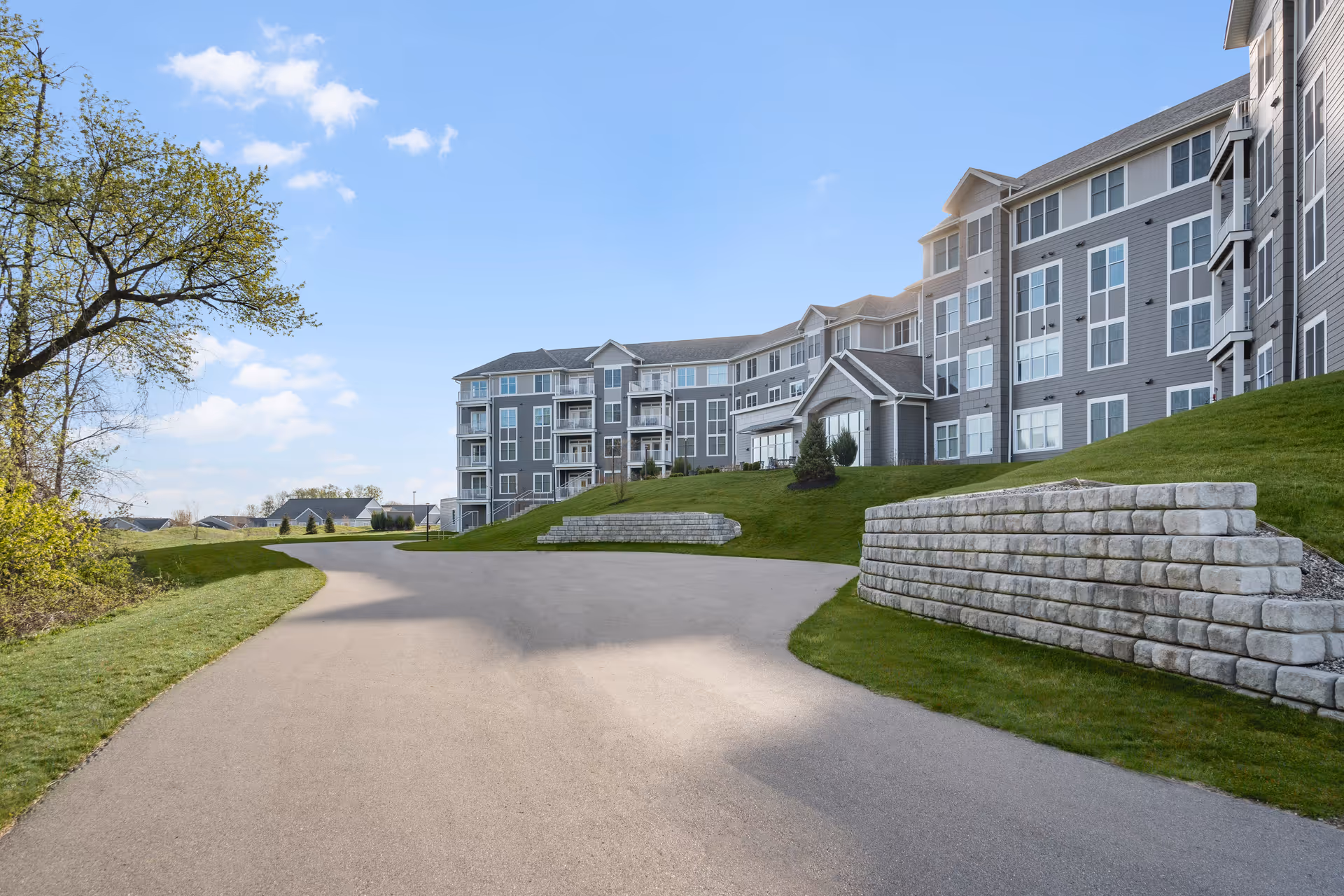 Exterior view of a large, multi-story senior living facility named Rivertown Ridge with gray siding and many windows. The building is situated on a grassy hill with stone retaining walls and a paved driveway leading up to it. Trees and a blue sky with some clouds are visible in the background.