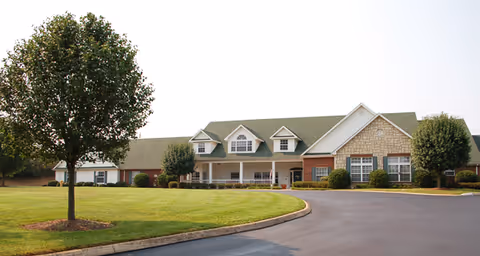 A single-story building with a green roof and a combination of brick and stone exterior walls, surrounded by neatly trimmed bushes and trees, with a curved driveway and well-maintained lawn in front.