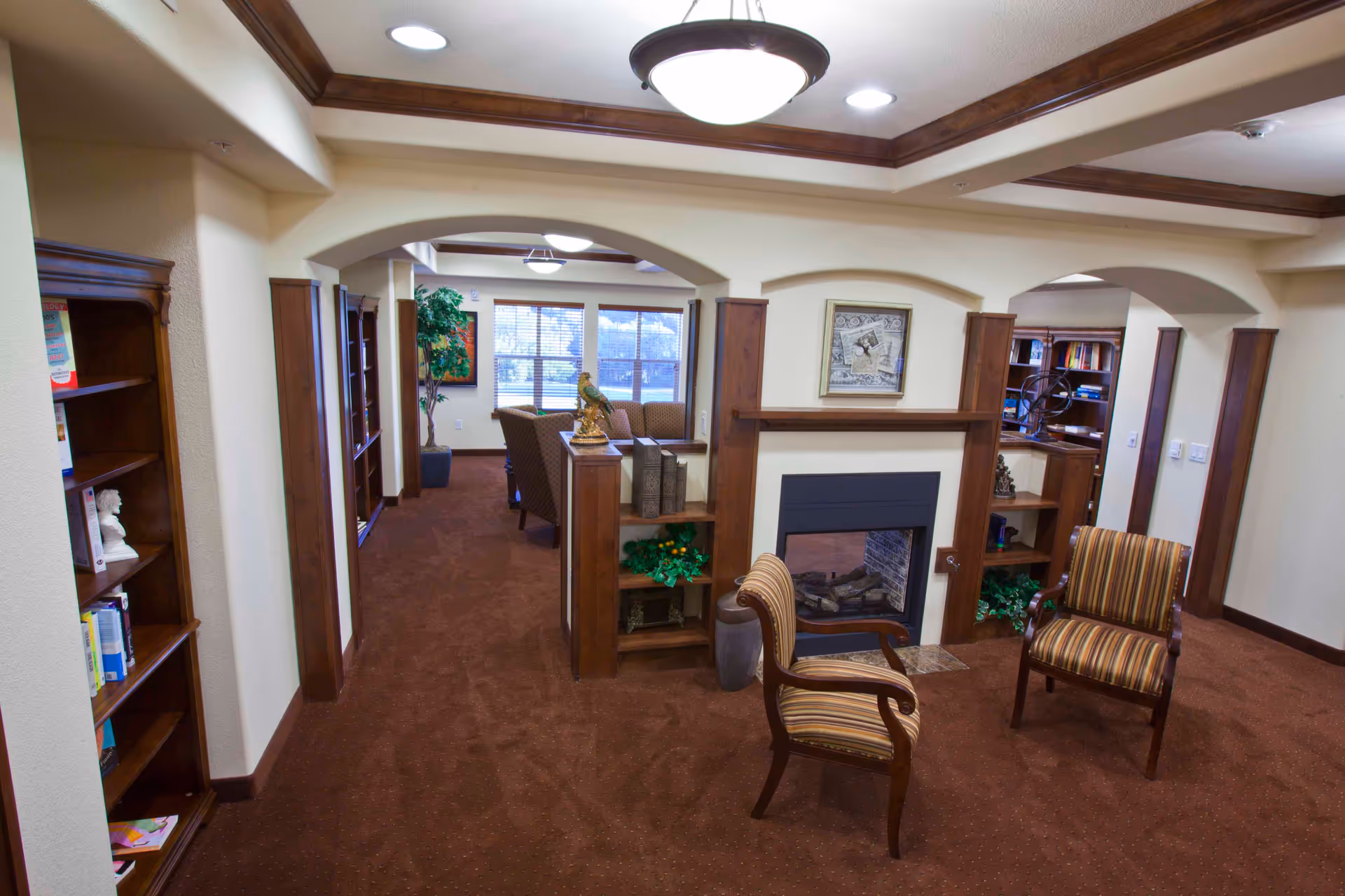 Interior view of a senior living facility lounge area with brown carpet, wooden trim, and beige walls. The room features a fireplace with built-in shelves on either side, two striped armchairs in front of the fireplace, and a seating area with chairs and a table near a window in the background. There are decorative plants and framed artwork on the walls.