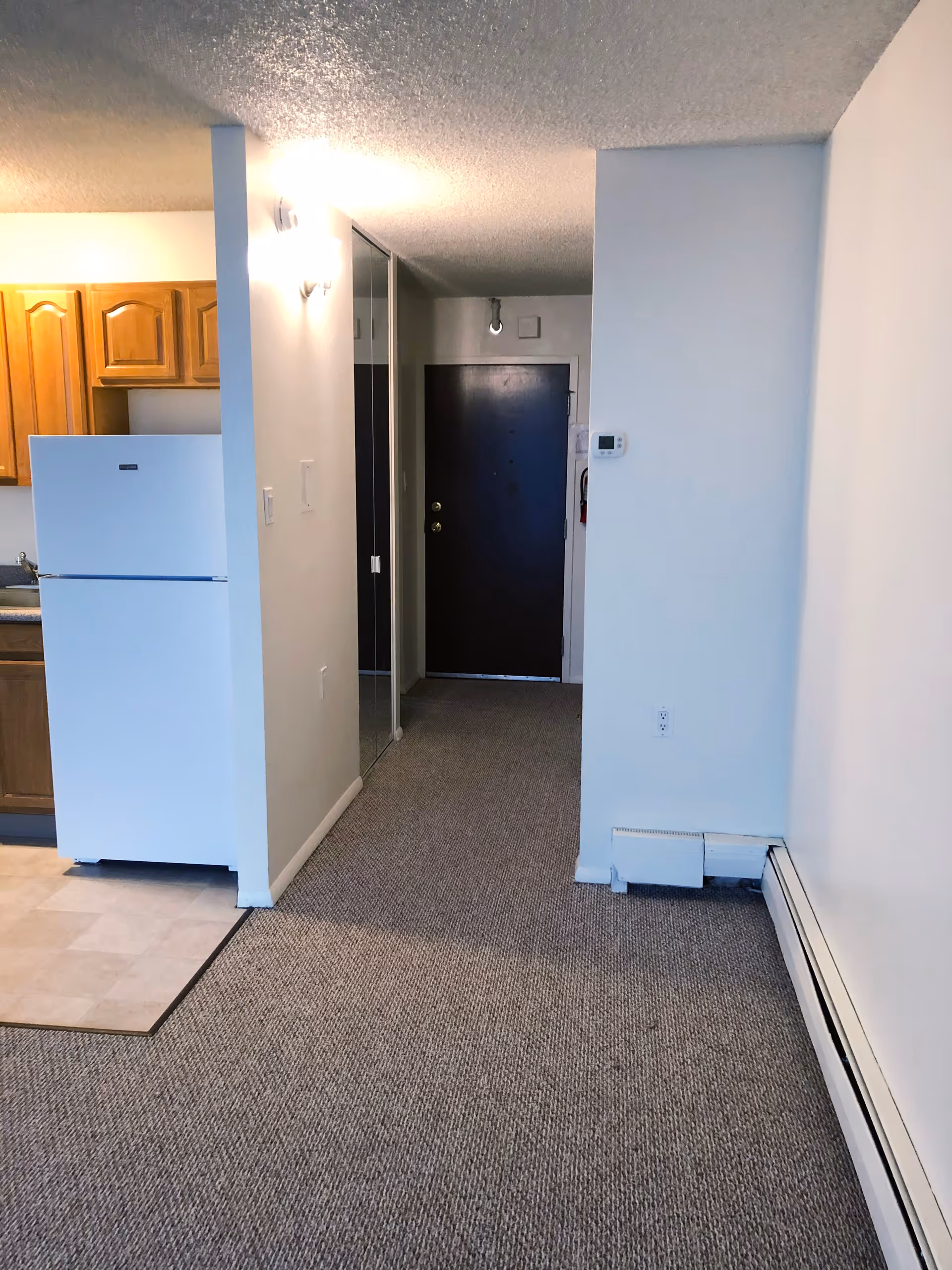 View of an apartment interior showing a hallway with carpeted floor leading to a dark front door. On the left side, there is a kitchen area with a white refrigerator and wooden cabinets. The walls are painted white, and there is a mirrored closet door along the hallway.