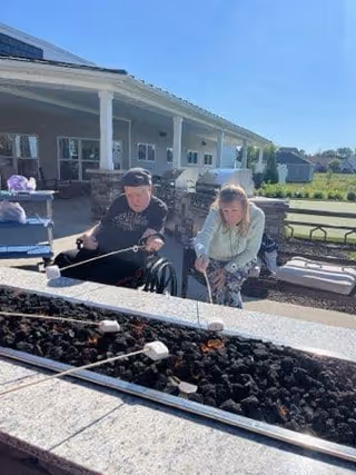 Two people sitting outdoors near a fire pit, roasting marshmallows on sticks. They are seated on benches with a covered patio and building in the background under a clear blue sky.