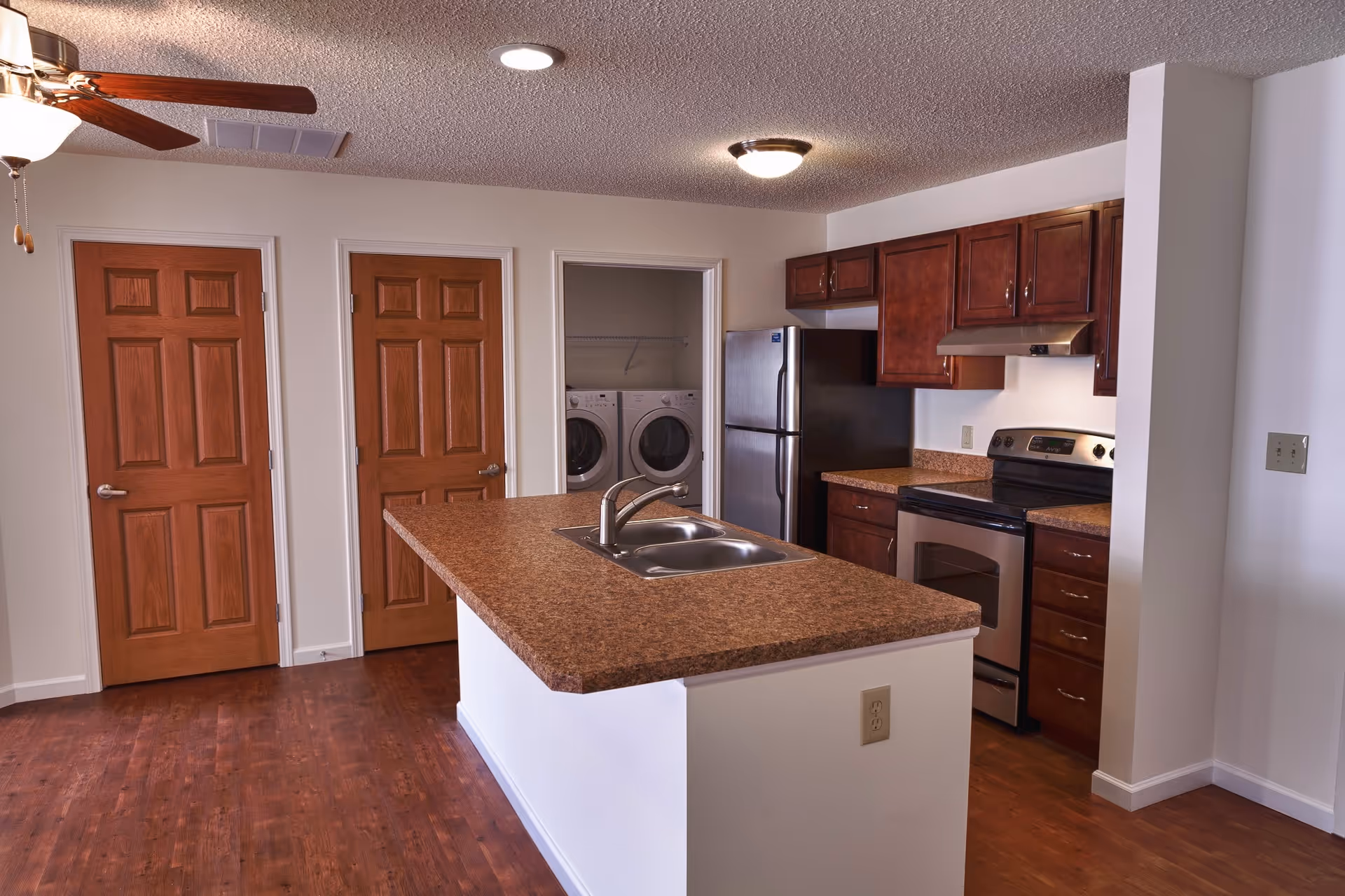 Interior view of a kitchen with a large island countertop featuring a double sink and faucet. The kitchen has wooden cabinets, a stainless steel refrigerator, and a stainless steel stove with an overhead range hood. There are two closed wooden doors and an open doorway revealing a laundry area with a washer and dryer. The floor is wooden, and the ceiling has a light fixture and a ceiling fan with lights.