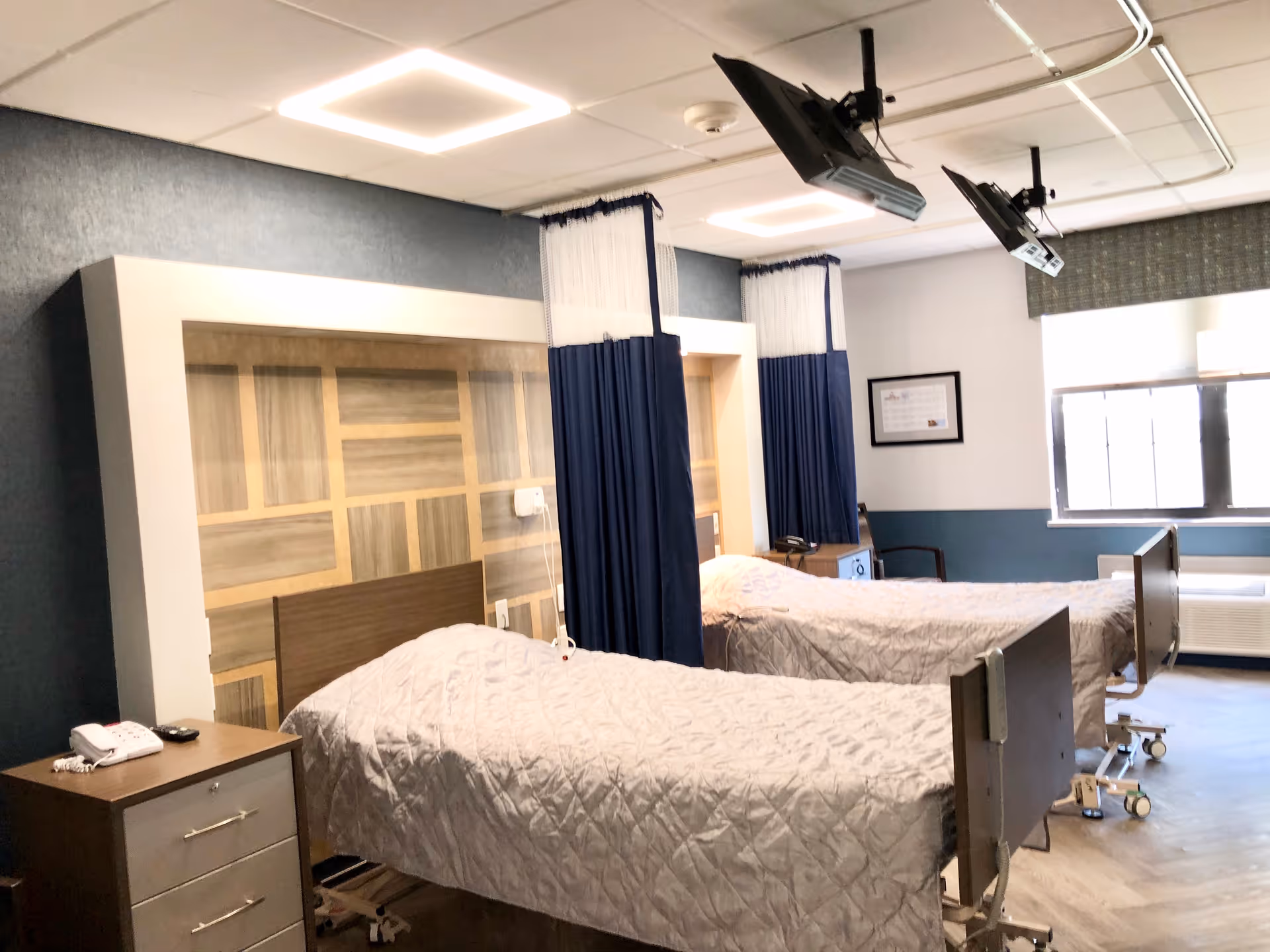 A room with two hospital-style beds separated by blue privacy curtains. Each bed has a beige quilted blanket. There is a wooden nightstand with a telephone and remote control next to one bed. The room has a large window with blinds, a framed picture on the wall, and two ceiling-mounted televisions positioned above the beds.