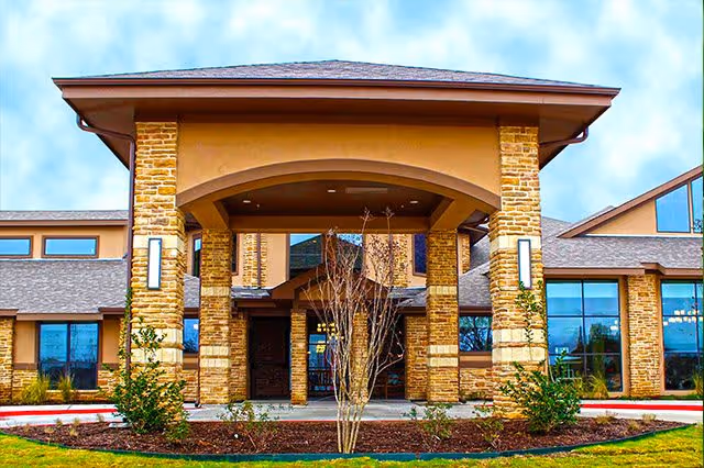 Front exterior view of Sterling Oaks Rehabilitation building with stone pillars, large windows, and a covered entrance under a partly cloudy sky.