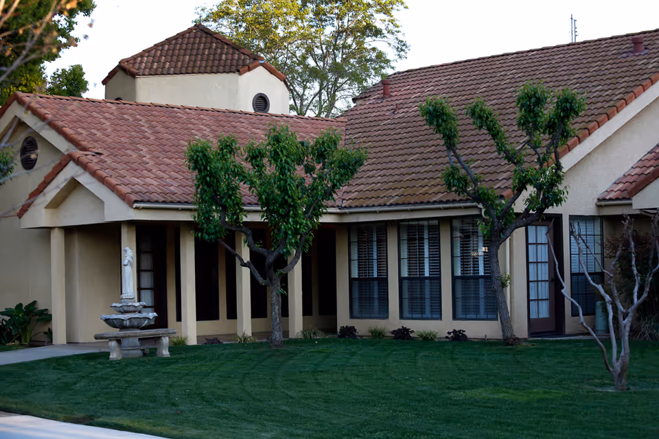 Exterior view of a single-story building with beige walls and a red tiled roof, surrounded by green grass and several small trees. There is a decorative fountain with a statue in front of the building.