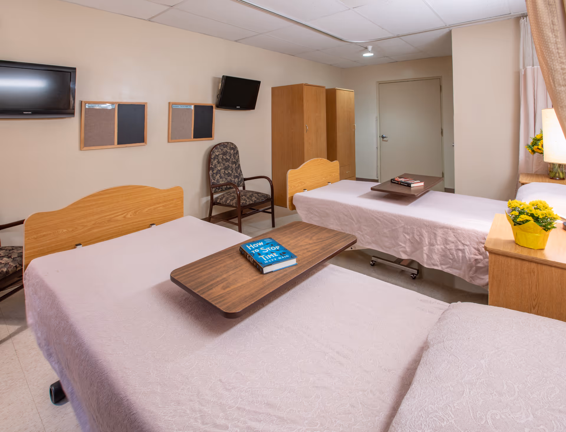 A shared patient room in a healthcare facility with two hospital beds covered in light pink bedspreads. Each bed has an overbed table with books on top. There are two wooden wardrobes against the wall, two patterned chairs, two wall-mounted televisions, and two bulletin boards. A small bedside table with a lamp and a yellow flower pot is visible near the window with curtains.