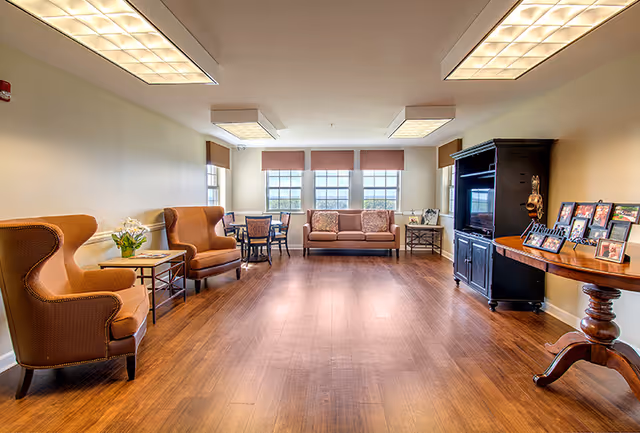 A bright and spacious living room with wooden flooring, featuring two brown upholstered armchairs on the left, a small table with a flower vase between them, a sofa with cushions against the far wall under three large windows with blinds, a round table with chairs near the windows, and a black entertainment center with a TV and framed photos on a wooden table on the right.