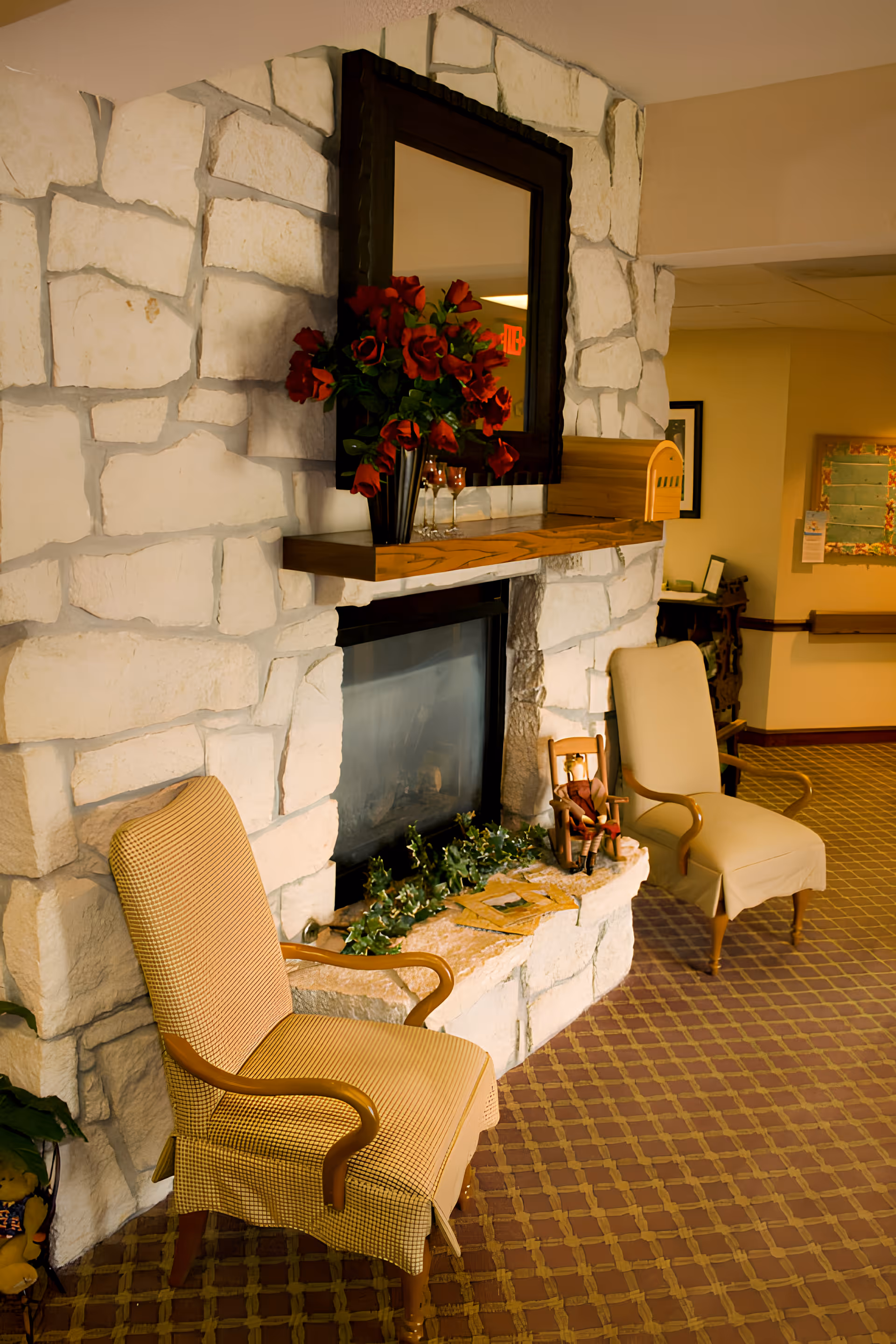 A cozy sitting area with two upholstered armchairs placed on a patterned carpet in front of a stone fireplace. Above the fireplace is a large framed mirror and a vase with red flowers. Decorative greenery and a small doll in a rocking chair are on the hearth. The background shows a hallway with framed pictures on the wall.
