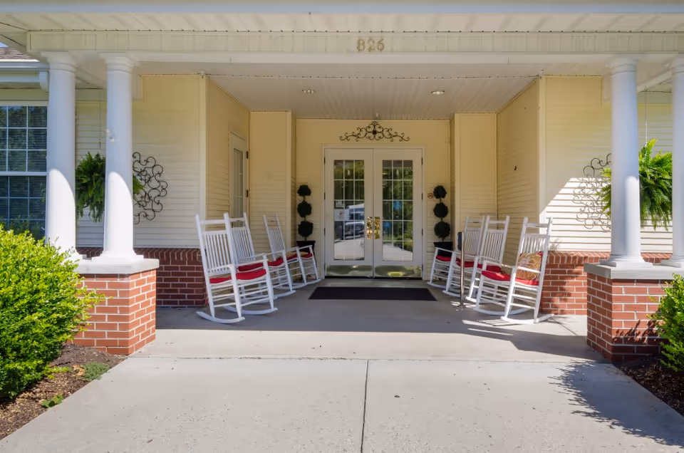 Entrance of a building with a covered porch featuring white columns and brick bases. There are white rocking chairs with red cushions arranged on both sides of the entrance door. Green plants and decorative wall hangings are visible on the walls beside the door.