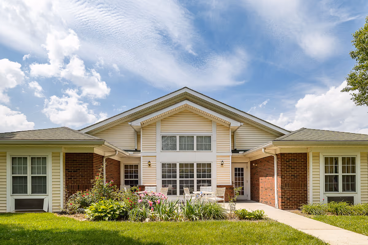 Front exterior of a single-story cottage with a central large window, patio seating, and landscaped flower beds under a blue sky.