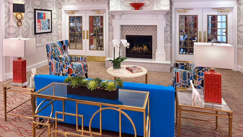 A cozy living room area featuring a blue sofa with a glass and gold metal console table behind it, two colorful patterned armchairs, a round coffee table with books and a potted orchid, and a white fireplace with a red decorative bowl on the mantel. The room has patterned carpet, wallpaper with circular designs, wall sconces, and double doors with glass panels.