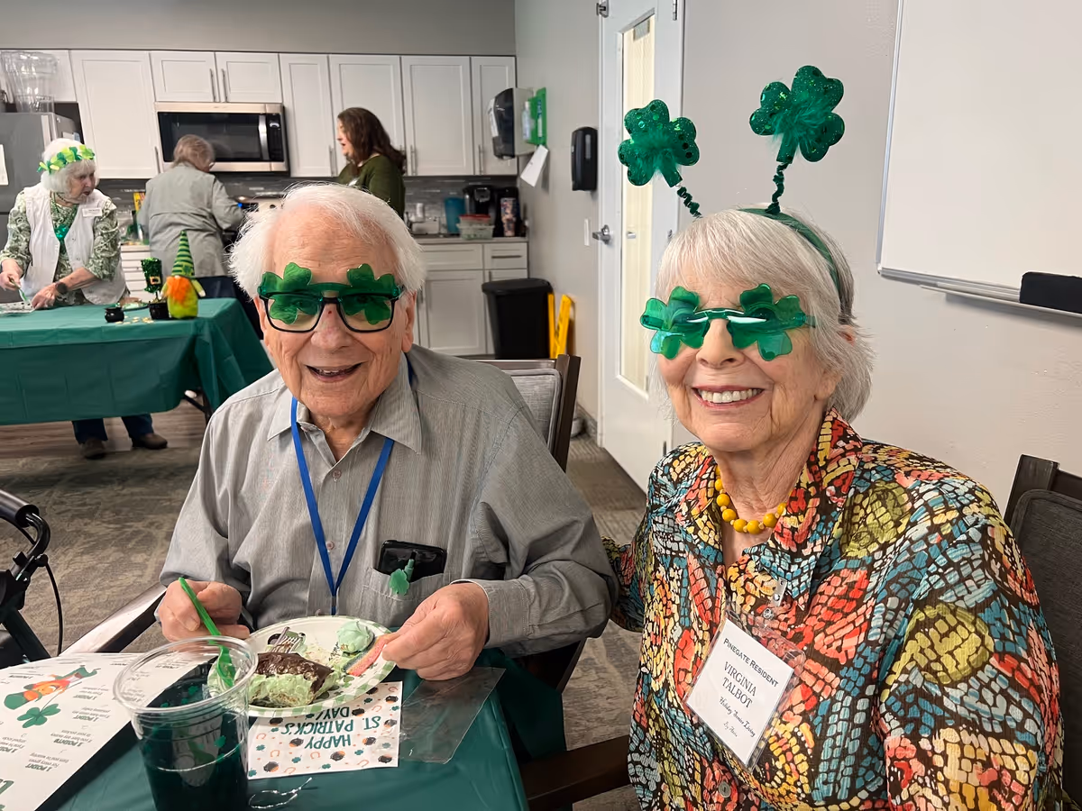 Two smiling elderly residents wearing shamrock-themed glasses and headbands seated at a table with cake and green drinks in a decorated indoor dining area.