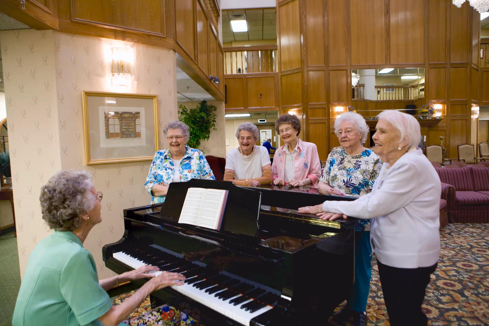 A group of elderly women gathered around a black grand piano in a spacious, wood-paneled common area. One woman is playing the piano while the others stand nearby, smiling and enjoying the music. The room features patterned carpet, upholstered chairs, and framed artwork on the walls.