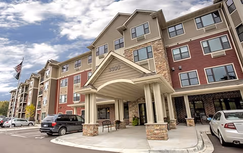 Exterior view of a multi-story senior living facility building with a covered entrance, several parked cars, and an American flag on a flagpole.