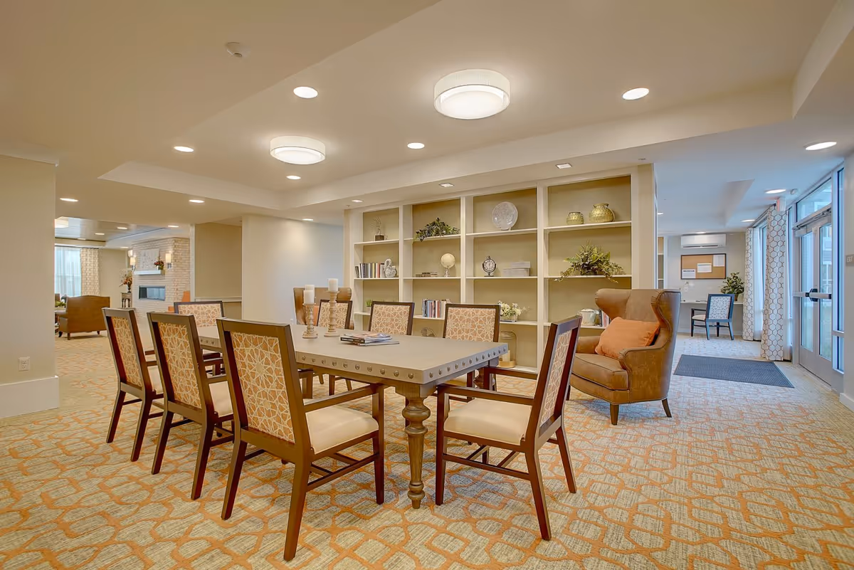 A well-lit interior common area with a rectangular table surrounded by eight chairs with patterned upholstery. Behind the table is a built-in shelving unit decorated with books, plants, and decorative items. To the right, there is a brown armchair with an orange cushion. The room has patterned carpet flooring and recessed ceiling lights. Large windows and glass doors are visible on the right side, allowing natural light to enter.
