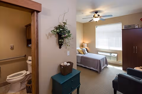 A small, carpeted bedroom with a single bed, dresser, armchair, and an open door showing an adjoining bathroom.