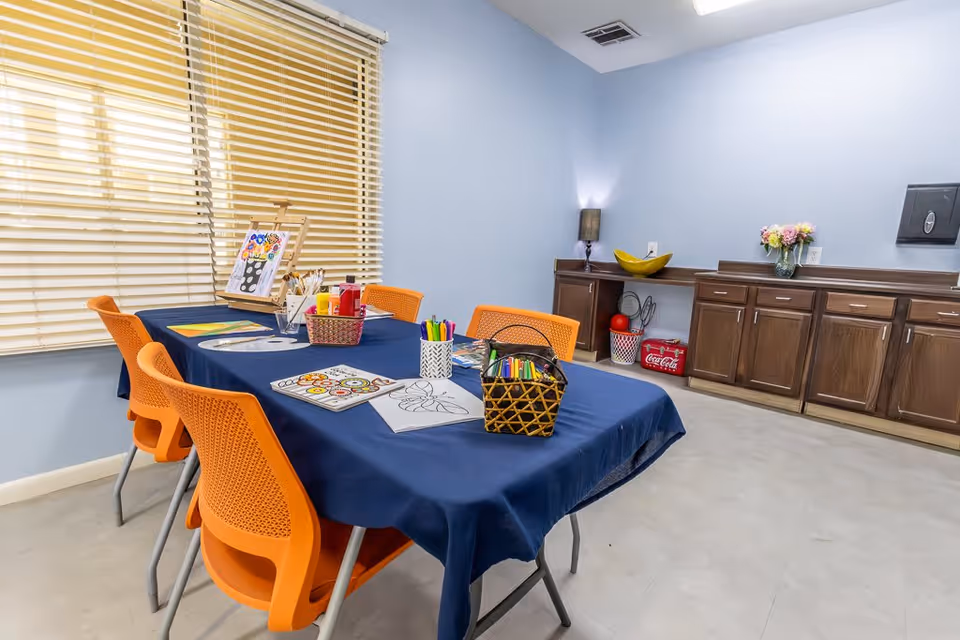 A bright activity room with a table covered by a blue tablecloth surrounded by six orange chairs. On the table are coloring books, colored markers, paint bottles, and an easel with a painting. The room has light blue walls, a window with closed blinds, and wooden cabinets along one wall with decorative items including a lamp, a bowl, flowers, and a Coca-Cola cooler.