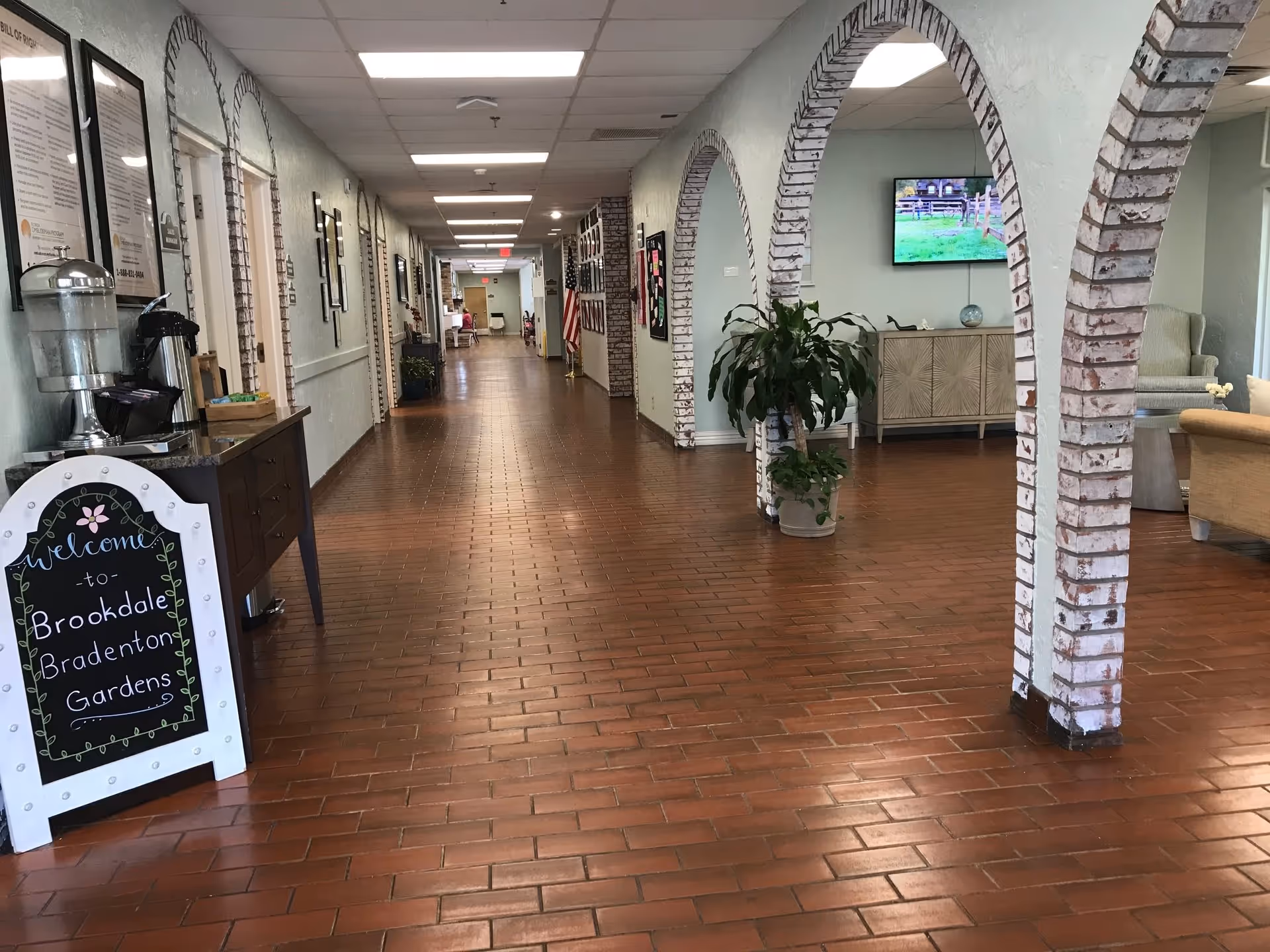 A long hallway in a senior living facility with brick archways on the right side leading to a seating area with chairs and a TV mounted on the wall. On the left side, there is a table with a water dispenser and coffee maker, and a decorative sign welcoming visitors to Brookdale Bradenton Gardens. The floor is covered with reddish-brown tiles and the walls are light blue with framed pictures and notices.