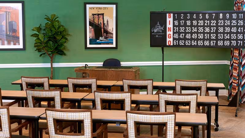 Activity room with rows of tables and chairs facing a podium and a large bingo board against a green wall.