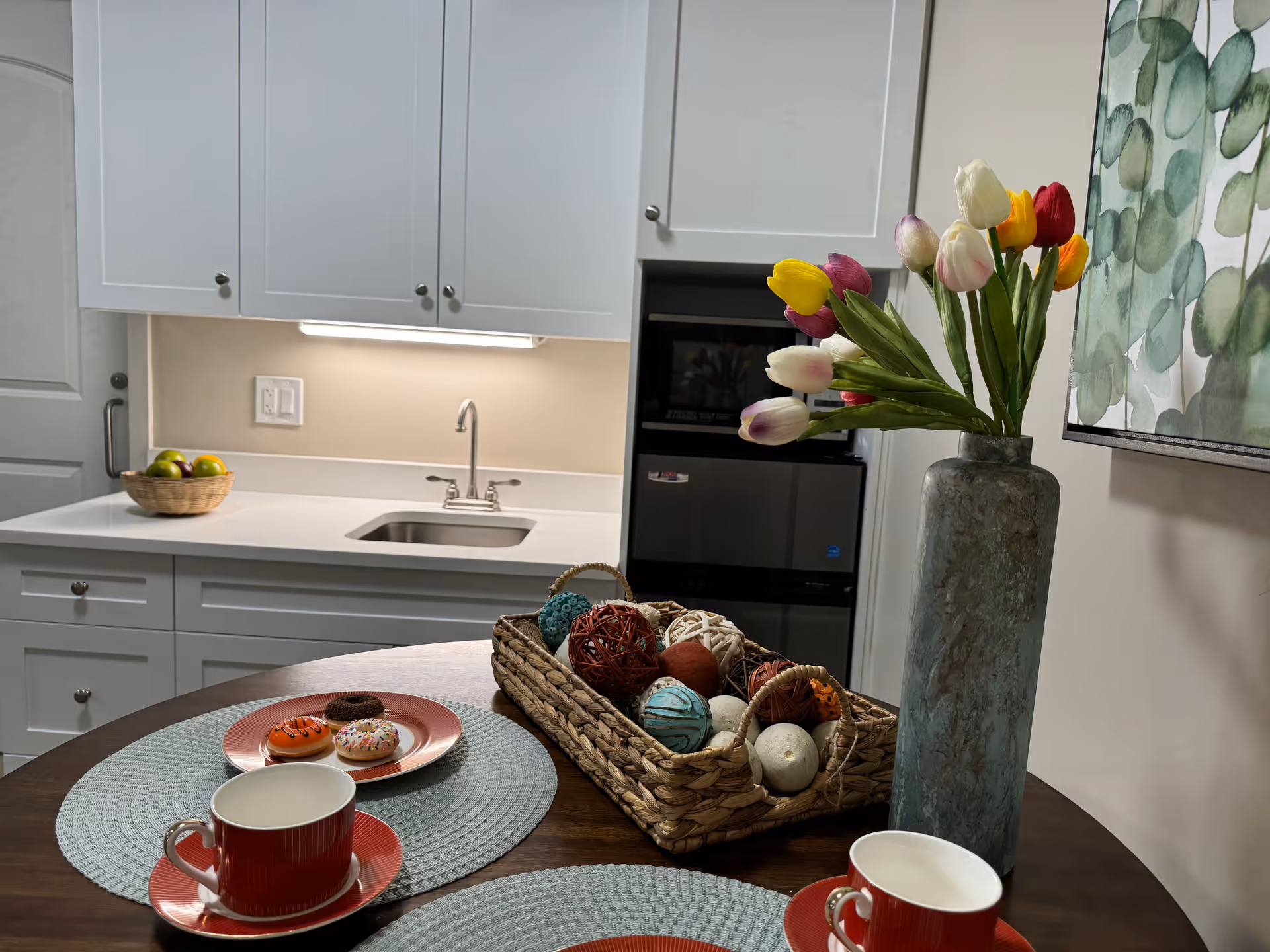 A small kitchen area with white cabinets, a sink, and a countertop. In the foreground, there is a round wooden table with two red cups and saucers, a plate with three donuts, a woven basket filled with decorative balls, and a tall vase holding colorful tulips. A framed botanical artwork is visible on the wall to the right.