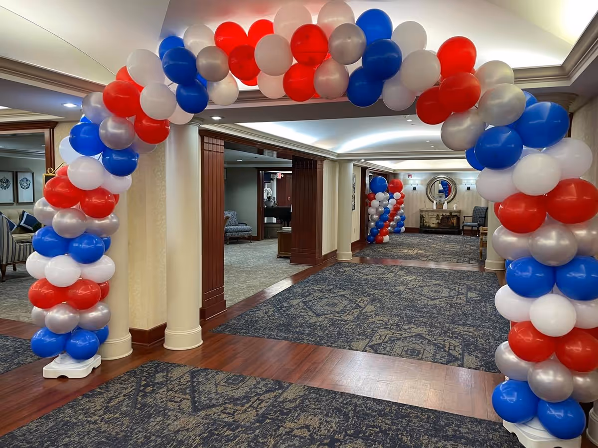 Interior hallway of a senior living facility decorated with an arch and columns of red, white, blue, and silver balloons. The hallway has patterned carpet, wood flooring accents, and beige walls with wooden trim. There are chairs and a mirror visible in the background.