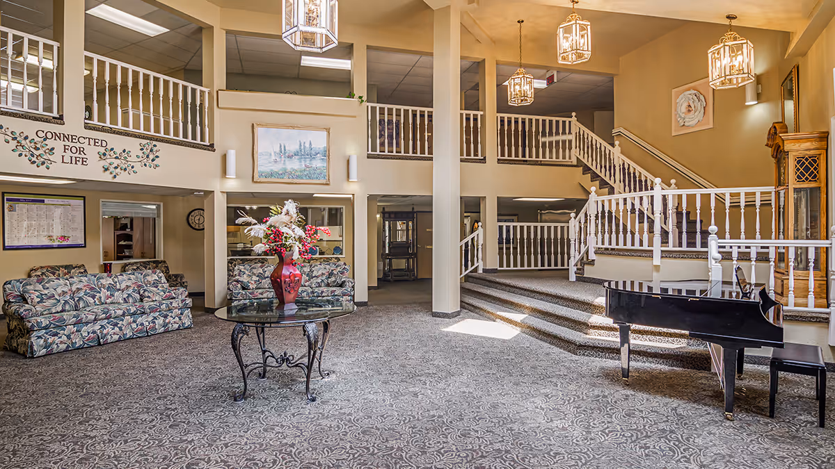 Spacious senior living facility common area with patterned carpet, floral couches, a glass table with a red vase of flowers, a black grand piano, and a staircase with white railings leading to an upper level. The walls are decorated with paintings and a sign that reads 'CONNECTED FOR LIFE'.