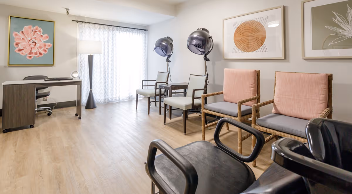 A bright room with wooden flooring featuring two vintage hair dryer chairs with overhead dryers, two wicker chairs with pink cushions, a desk with an office chair, a tall floor lamp, and three framed artworks on the walls.