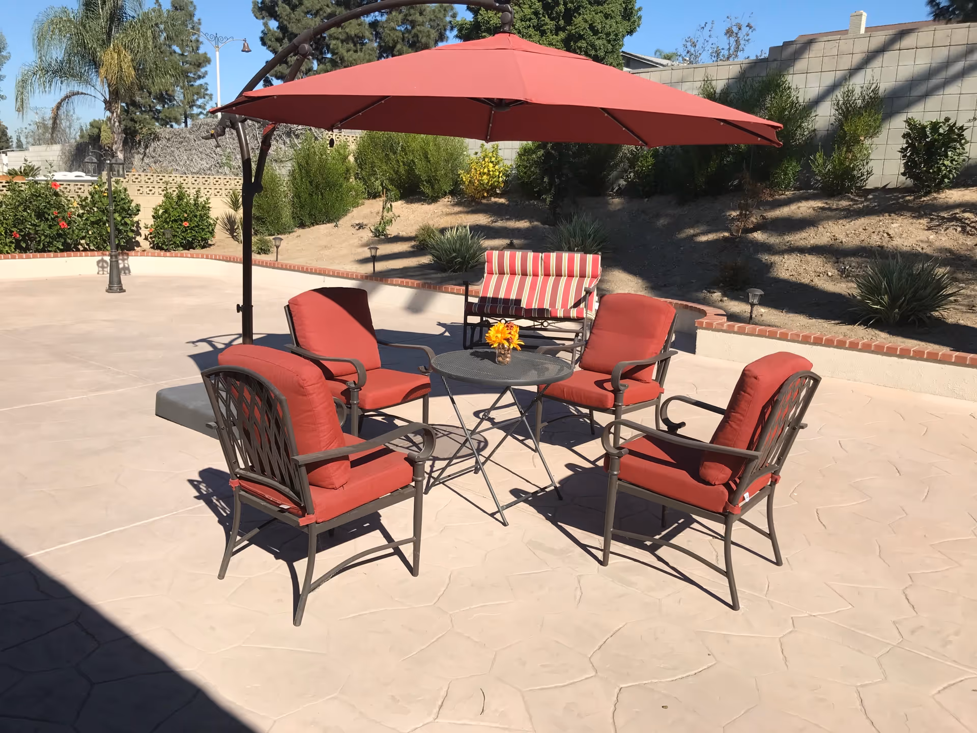 Outdoor patio area with four red cushioned metal chairs arranged around a small round metal table with a small flower arrangement on top. A large red umbrella provides shade over the seating area. In the background, there is a striped cushioned bench and a garden with various plants and shrubs against a block wall.