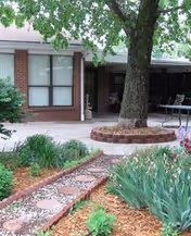 Brick building exterior of a senior living facility with a tree, garden, and walkway.