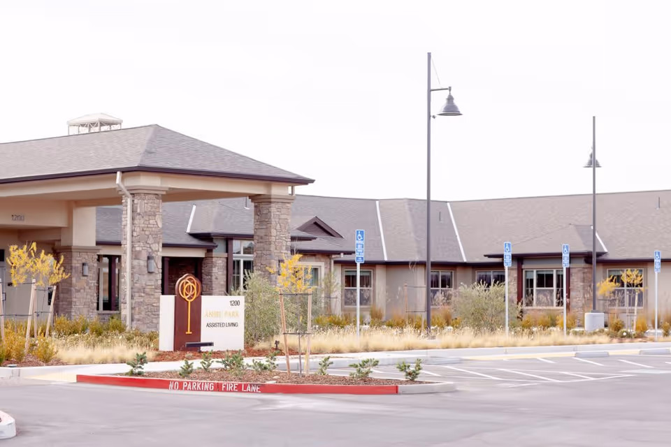 Exterior view of Ansel Park Assisted Living & Memory Care facility showing the entrance with stone pillars, a sign with the facility name, and a parking lot with designated handicapped parking spaces and street lamps.