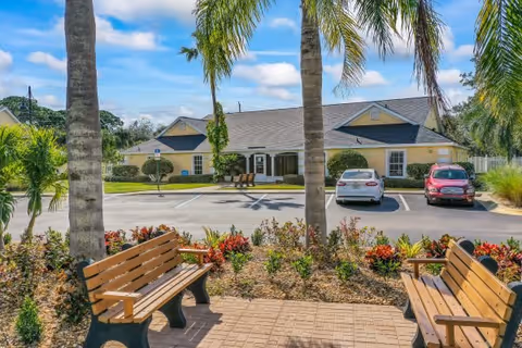 Outdoor view of Palm Cottages of Rockledge showing a single-story building with a gray roof and yellow walls. In the foreground, there are two wooden benches facing each other with a landscaped area of plants and palm trees between them. A parking lot with two cars is visible in front of the building under a partly cloudy sky.