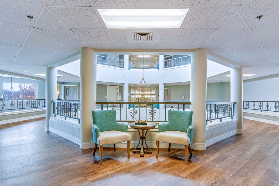 Bright open indoor seating area with two green armchairs and a small table overlooking a central atrium with railings and a chandelier.