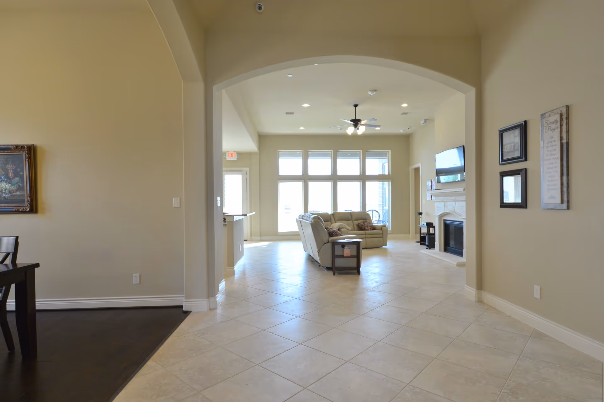 View through an arched doorway into a bright living room with large windows, a ceiling fan with lights, a mounted flat-screen TV above a fireplace, and a beige sofa set with a side table. The floor is tiled, and the walls are painted light beige. Part of a dining area with dark wood flooring and a framed painting is visible on the left.