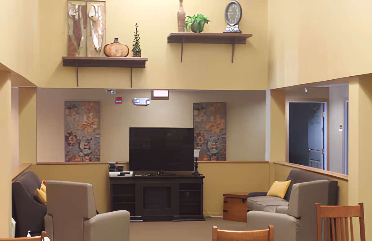 A cozy living room area with beige walls and a high ceiling. The room features a flat-screen TV on a dark wooden stand, two abstract paintings on the wall behind the TV, and two floating shelves above with decorative items including a plant, a small sculpture, and vases. There are two gray armchairs and a gray sofa with yellow cushions arranged around the TV. A wooden chair and a small wooden chest are also visible.