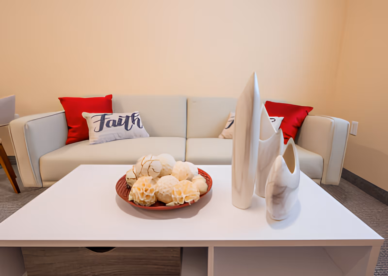 Bright living room with a white sofa, red accent pillows, and a white coffee table holding decorative vases and a bowl of decorative spheres.