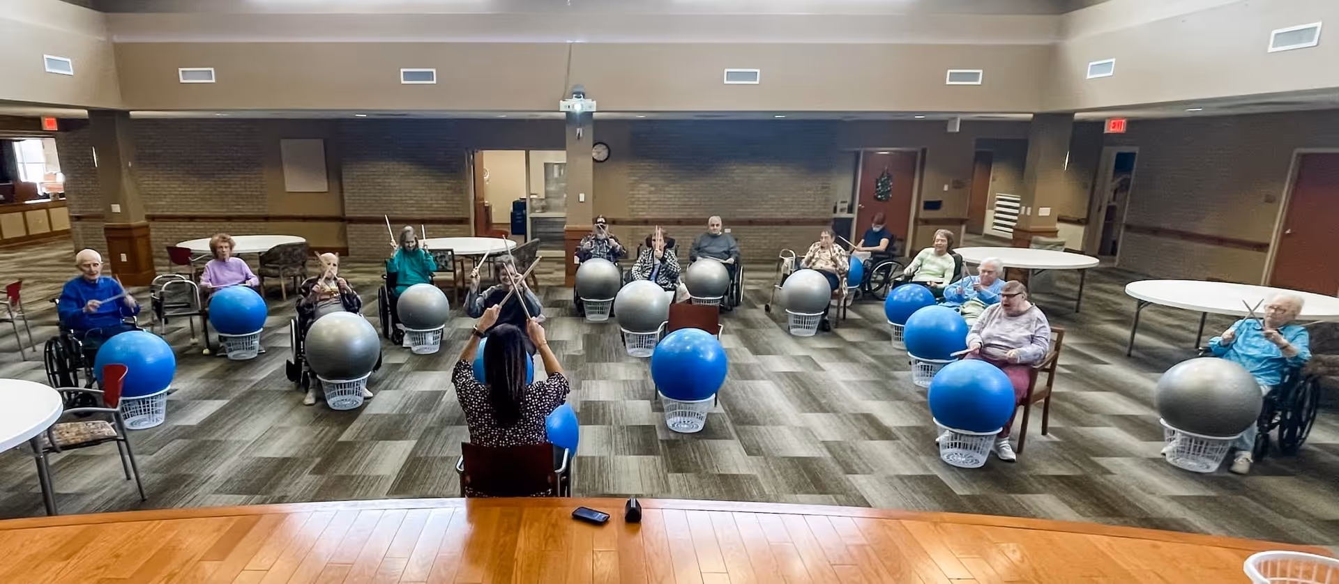 A group of elderly individuals seated in a spacious room participating in a drumming exercise led by an instructor. Each participant has a large exercise ball placed on a laundry basket in front of them and is holding drumsticks. The instructor is seated facing the group with drumsticks raised. The room has carpeted flooring, round tables, and a high ceiling with vents.