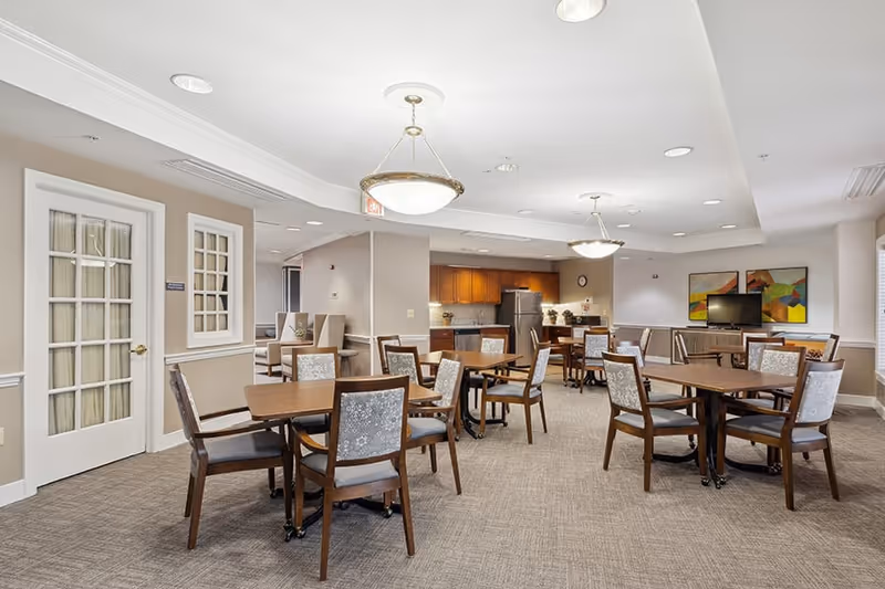 A spacious dining area in an assisted living facility featuring several wooden tables with cushioned chairs arranged neatly. The room has beige walls, carpeted floors, and ceiling lights. In the background, there is a kitchen area with wooden cabinets, a refrigerator, and a countertop. A TV is mounted on the far wall with colorful abstract paintings beside it.