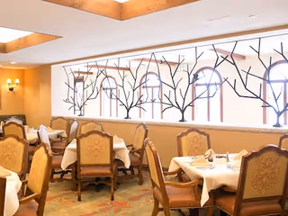 Dining room with several tables covered with white tablecloths and set with napkins and silverware. The chairs have ornate wooden frames and upholstered backs and seats. The room has large windows with decorative black tree branch designs and warm lighting from wall sconces and ceiling beams.