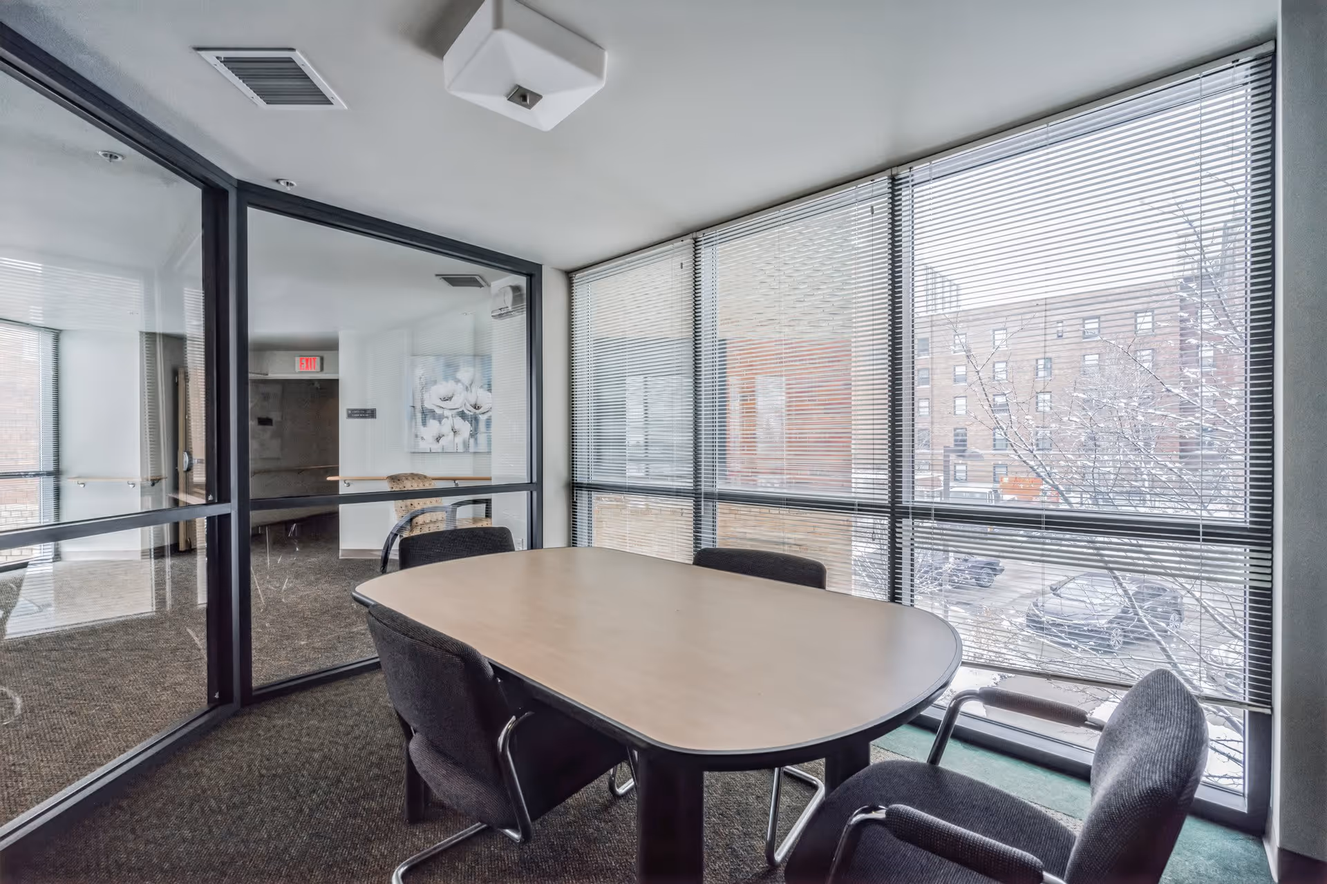 Glass-walled meeting room with a rectangular table and four chairs beside large windows overlooking a snowy parking lot.