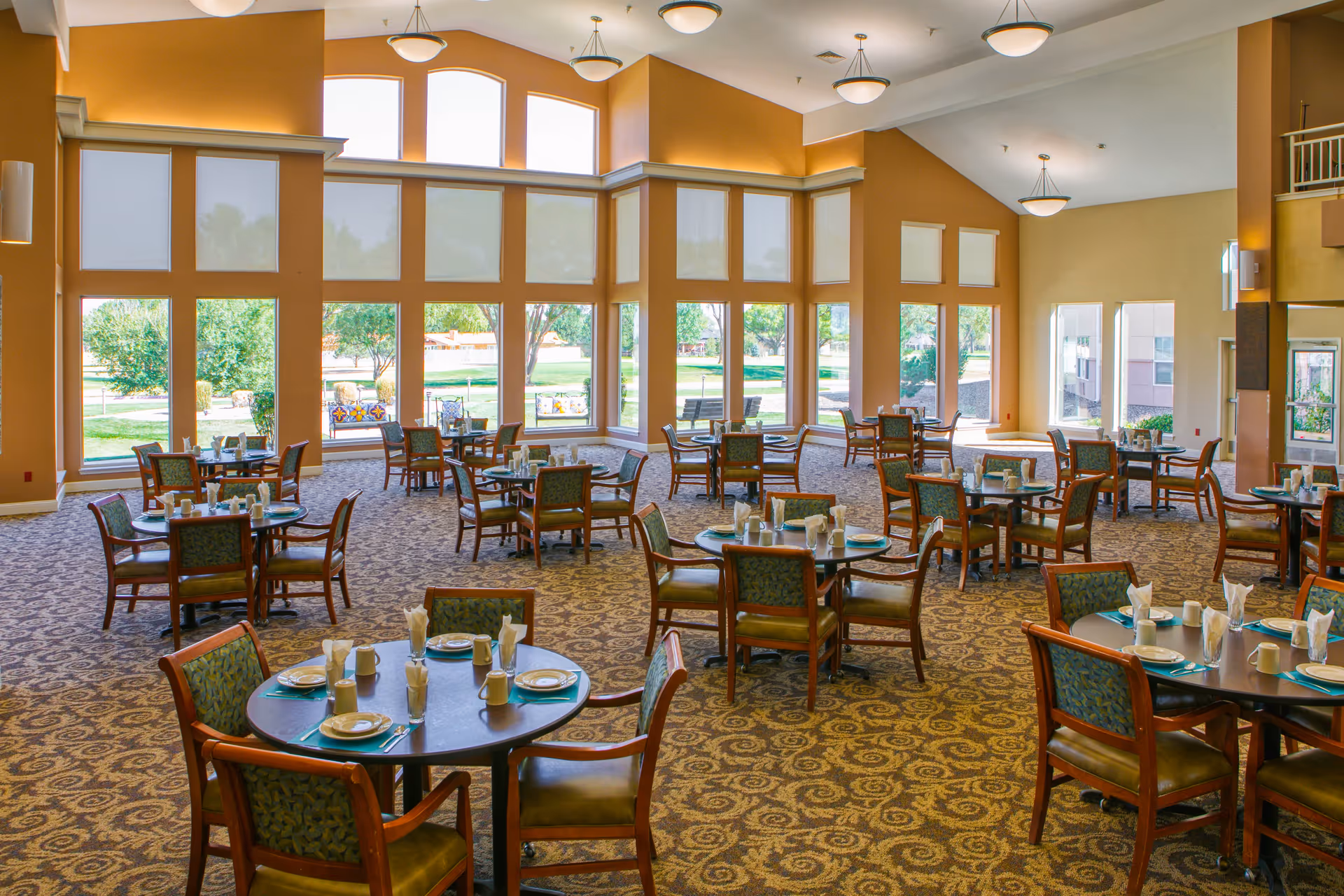 A spacious dining room with multiple round tables set with plates, cups, and napkins. The room features large windows letting in natural light and offering views of a green outdoor area with trees and benches. The walls are painted in warm tones and the ceiling has several hanging light fixtures.